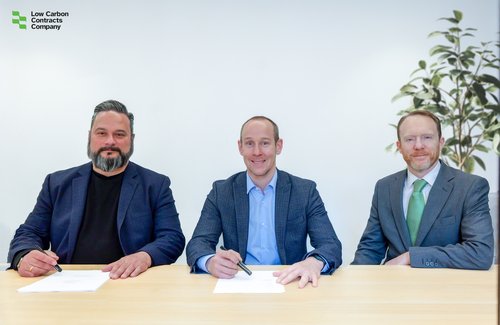 Three individuals seated at a table, each wearing business attire, holding pens and positioned in front of a document.