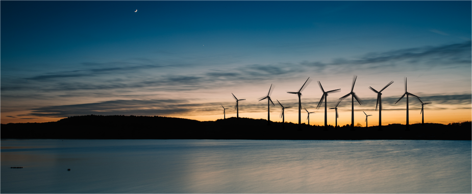 Water in the foreground with an array of windmills in the background, as the sun sets behind it