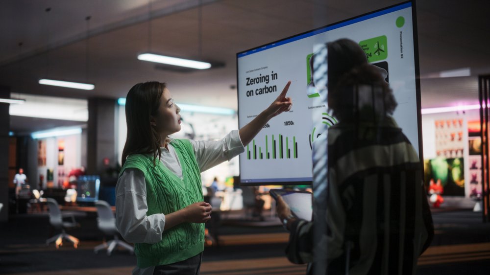 A woman points at a large monitor, while another woman listens attentively. 